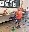 Child standing in a classroom next to a desk with one arm raised.