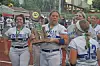 Softball players in white uniforms hold a trophy while standing together outdoors.