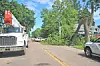 A utility truck and a fallen tree lie across a residential street lined with houses and trees.