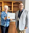 Two people stand in front of wooden shelves holding a framed certificate.