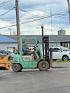 Green forklift driving past parked cars and warehouses on a street.