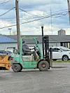 Green forklift driving past parked cars and warehouses on a street.