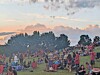 Large crowd standing and sitting on a grassy field at sunset with smoke in the air and trees behind them.