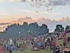 Large crowd standing and sitting on a grassy field at sunset with smoke in the air and trees behind them.