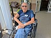 An elderly man sits in a wheelchair on a porch outside a house.