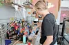 Person standing behind a cafe counter preparing a drink among bottles, cups and equipment.