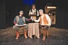 Three performers seated on a small stage with a small table, dark curtains, and rustic background props.