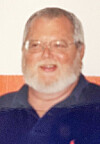 An older man wearing glasses and a navy shirt poses indoors against a plain background.
