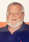An older man wearing glasses and a navy shirt poses indoors against a plain background.