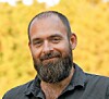 Bearded man in a dark shirt posing outdoors against a blurred yellow background.