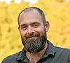 Bearded man in a dark shirt posing outdoors against a blurred yellow background.