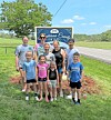 A group of people posing on grass beside a Harmony County Home sign near a roadside.