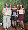 Five women standing together indoors, each holding framed certificates or awards in front of a green wall.