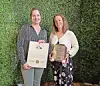 Two women posing with framed award plaques against a green plant wall.