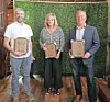 Three people stand indoors holding framed award plaques in front of a green hedge wall.