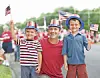 Children and an adult smiling and waving small American flags at an outdoor event.