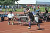 Runner in dark uniform lunges forward on a track near starting equipment and seated officials.