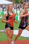 Two female runners compete on a track with a blurred crowd behind them.