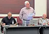 A man stands reading from paper at a meeting table with two seated men, a laptop, and water bottles.