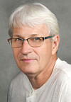 Close-up portrait of a man with white hair, glasses, and a light shirt on a gray background.