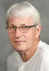Close-up portrait of a man with white hair, glasses, and a light shirt on a gray background.