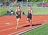 Two runners competing on an outdoor track with a few spectators and parked cars behind a fence.