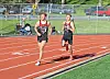 Two runners competing on an outdoor track with a few spectators and parked cars behind a fence.