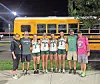 A group of softball players and coaches stand in front of a yellow school bus at night.