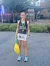 A woman in running gear holds a race bib, medal, and yellow bag outside a building.