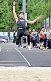 An athlete is captured midair on an outdoor track with spectators and trees in the background.
