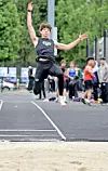 An athlete is captured midair on an outdoor track with spectators and trees in the background.