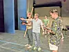 People on a stage rehearsing, one reading from a script while others stand near a long wooden prop.