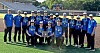 Youth baseball team posing on a baseball field with a trophy in front of stadium seats.