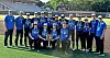 Youth baseball team posing on a baseball field with a trophy in front of stadium seats.