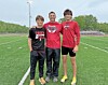 Three young men standing on a green sports field beside a running track and empty bleachers.