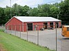 Red barn-style building behind a chain-link fence with a gravel lot and an orange vehicle at right.