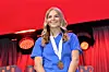 Athlete in a blue jersey wearing a medal stands on a podium against a red-lit stage backdrop.