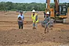 Three workers in hard hats stand on a dirt construction site beside an excavator.