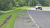 A car drives on a curving road beside a grassy shoulder and trees.