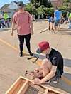 People working on wooden frames and tools in an outdoor paved area on a sunny day.