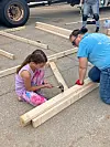 A child and an adult kneel beside lumber pieces, using tools to build a wooden frame outdoors.