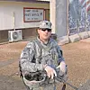 A soldier in camouflage uniform stands holding gear outside a post office building.