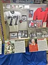 Glass display case with football jerseys, photos, articles, and memorabilia arranged on shelves.