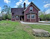 Red farmhouse with white trim, front porch, driveway and grassy yard under a blue sky.