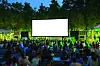 Audience seated outdoors in front of a large blank screen in a wooded setting at dusk.