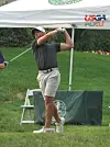 Golfer in cap and golf attire swinging a club on a tee box near a tent with USGA branding.