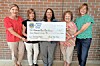 Five women standing together holding a large ceremonial check in front of a brick wall.