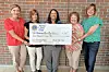 Five women standing together holding a large ceremonial check in front of a brick wall.