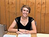 Smiling woman seated at a table with papers in front of a wooden wall.