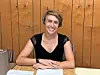 Smiling woman seated at a table with papers in front of a wooden wall.